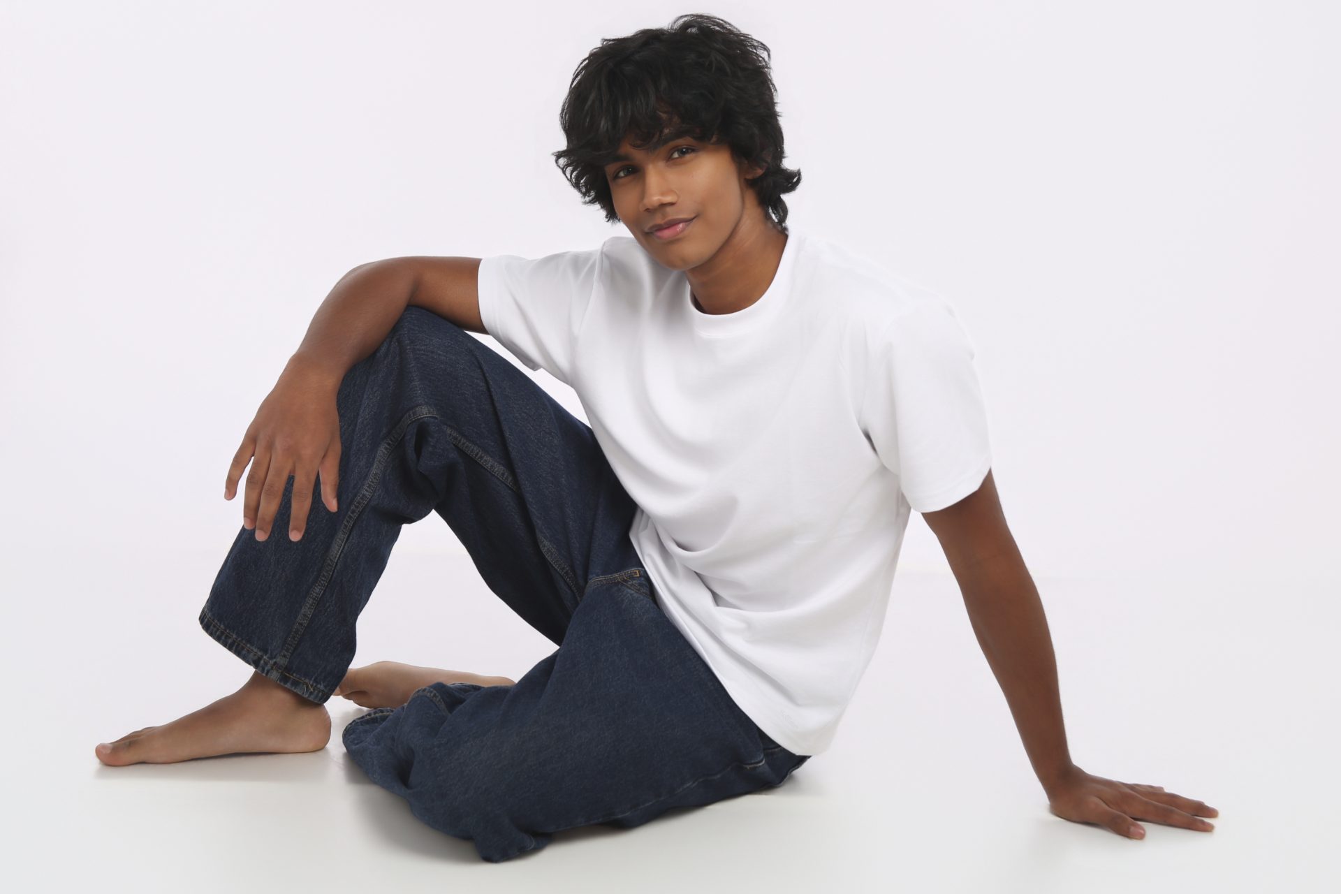 Shabs seated on a white studio floor in a white tee and denim